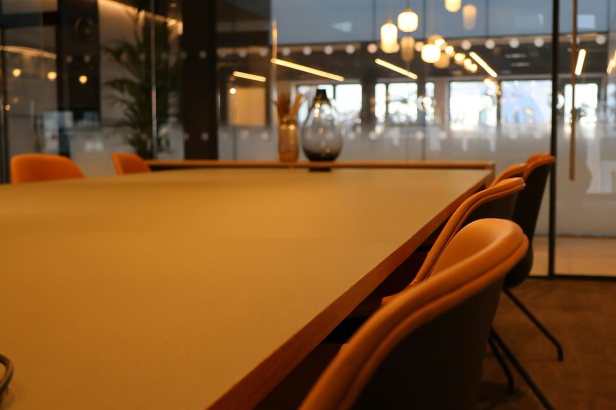 An empty meeting room with a long table and orange chairs, lights glowing in the background