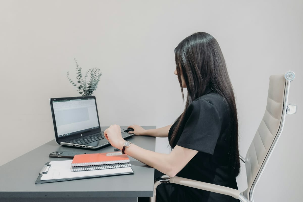 A woman working alone at a laptop at a small desk, with a notebook and a clipboard beside her
