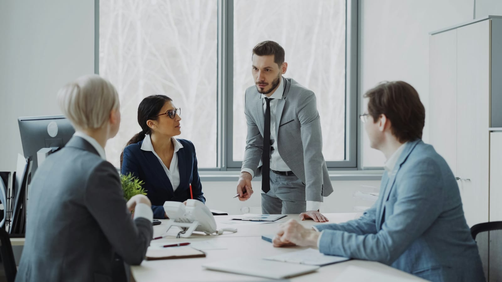 Four people at a meeting table in a bright office, one standing and leading the discussion