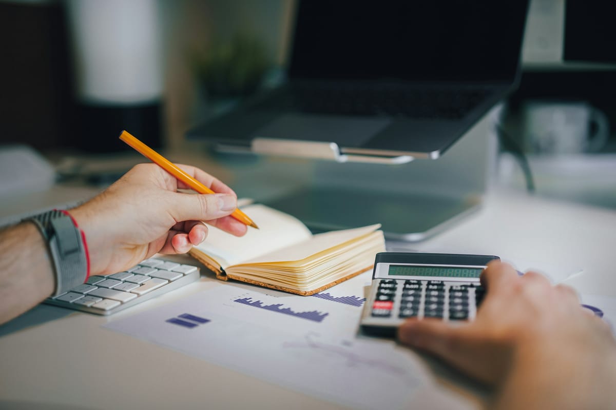 A person writing budget notes in a notebook with a pencil while holding a calculator, a laptop behind
