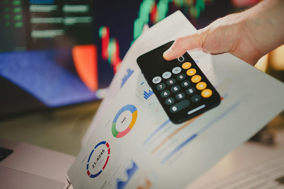 A hand holding a phone calculator over printed charts, with trading screens blurred in the background