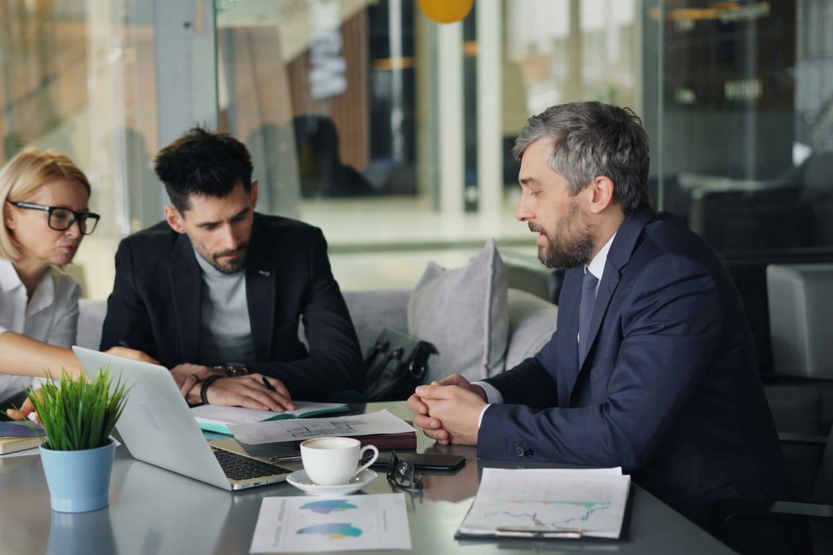A finance advisor in a suit talking with two colleagues over a laptop and printed charts