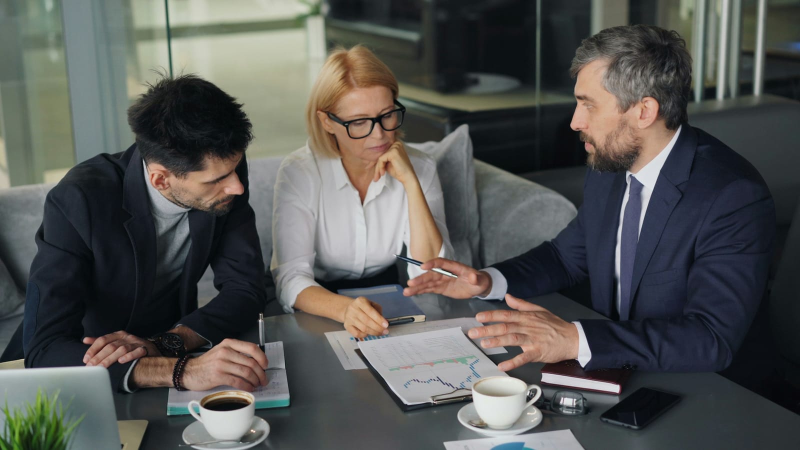 Three people in business clothes at a meeting table, going through a printed financial chart over coffee