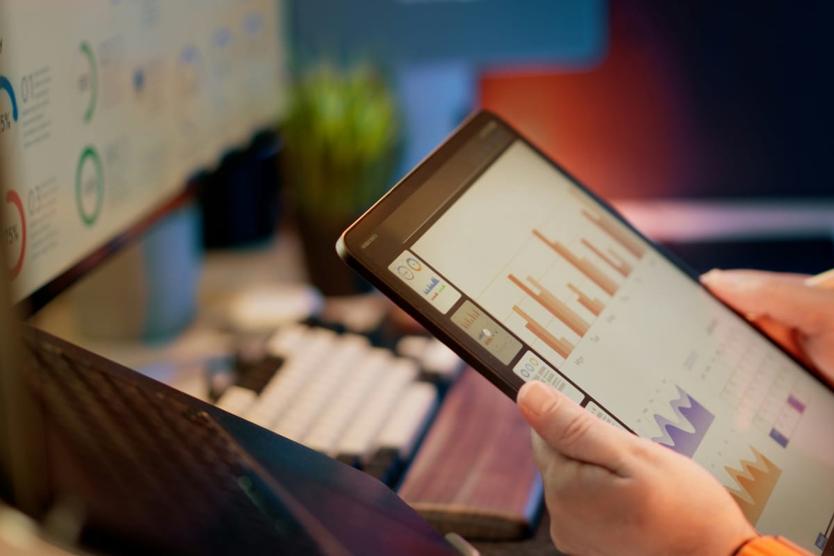 Hands holding a tablet that shows a bar chart report, with a keyboard and a monitor behind